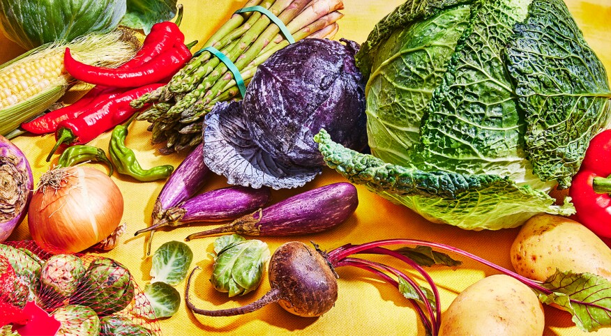 A variety of vegetables on yellow surface, including peppers, corn, eggplant, onions, brussels sprouts, cabbage, potatoes