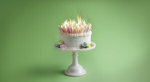 white birthday cake on a cake stand with a top full of lit candles.