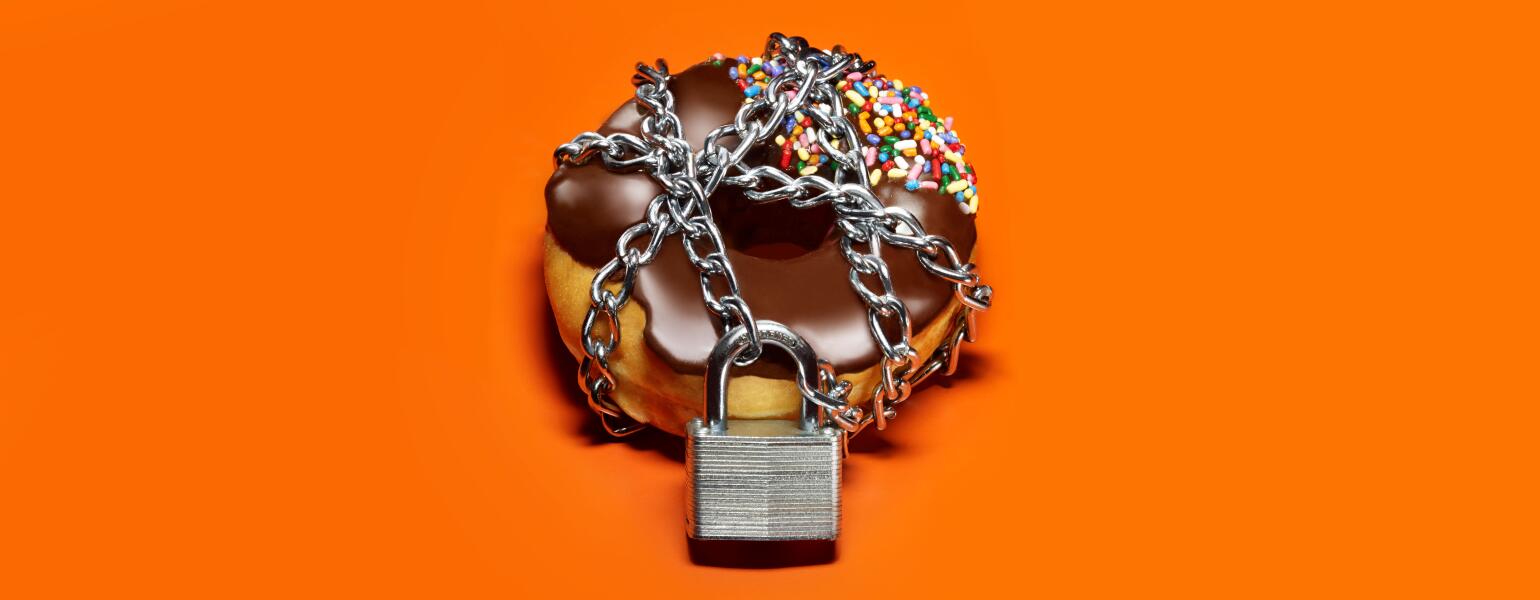A chocolate frosted donut on an orange background with a chain and lock around it.