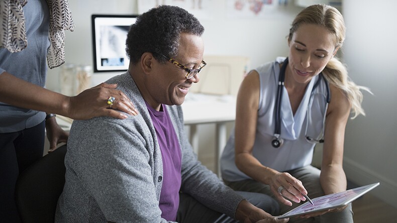 A blonde female doctor shows an older black man a tablet during a medical visit.