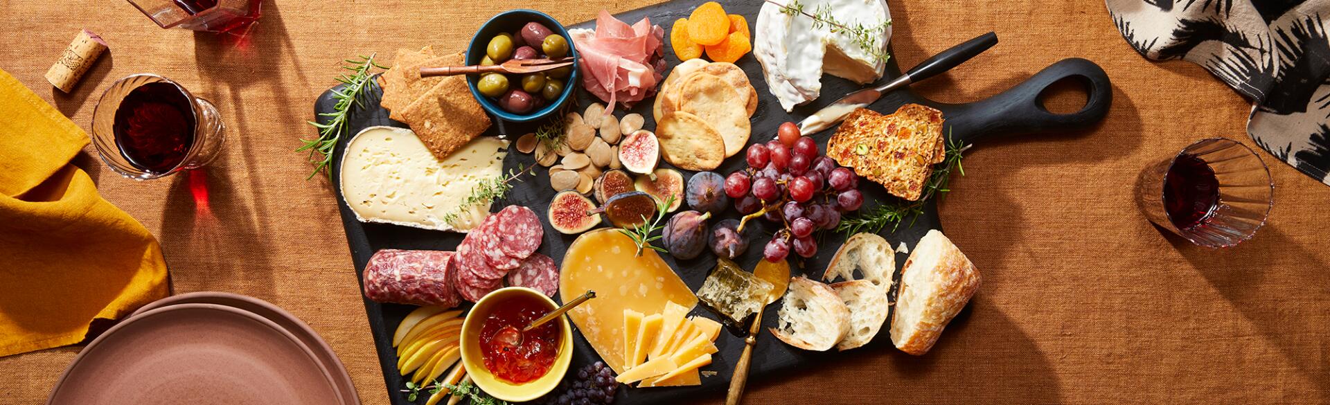 Charcuterie board with a display of cheese and meats on a rust colored table cloth, red wine, plates and napkins
