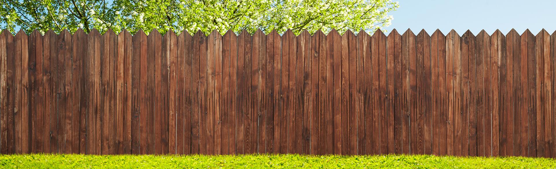 wood stained picket fence in green yard with tree behind it