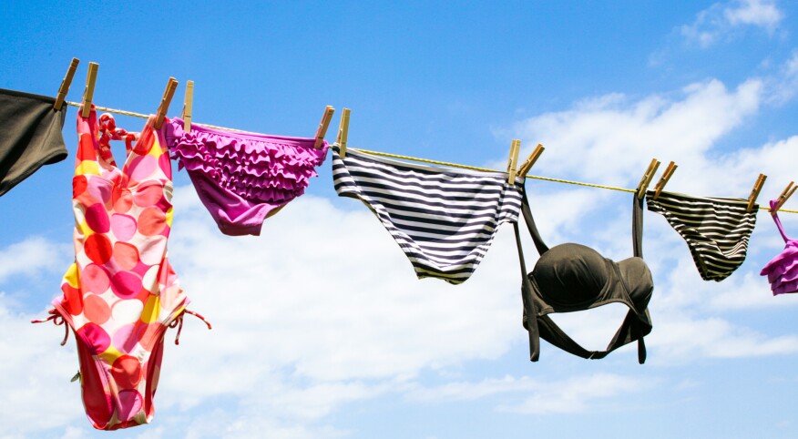 A close line with a blue sky and white clouds behind it. Bathing suits from left to right, moss green bottoms, a pink, orange, yellow and white polka dot one piece, purple ruffle bikini bottoms, black and white stripped bikini bottoms, a black bikini top, another black and white stripped bikini bottom, and a purple ruffle bikini top.