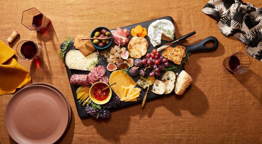 Charcuterie board with a display of cheese and meats on a rust colored table cloth, red wine, plates and napkins