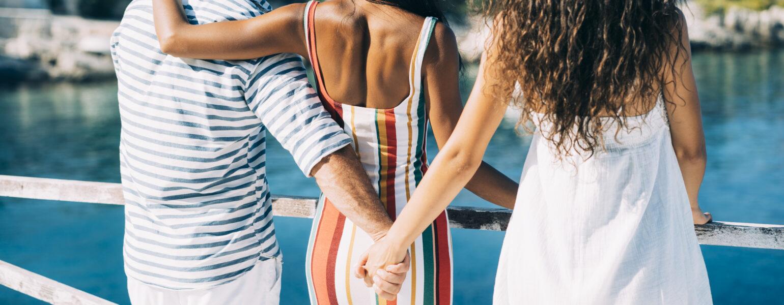 A man stands to the left of two women. While the woman in the middle has her arm around the man, he is holding the hand of the other woman behind her back.