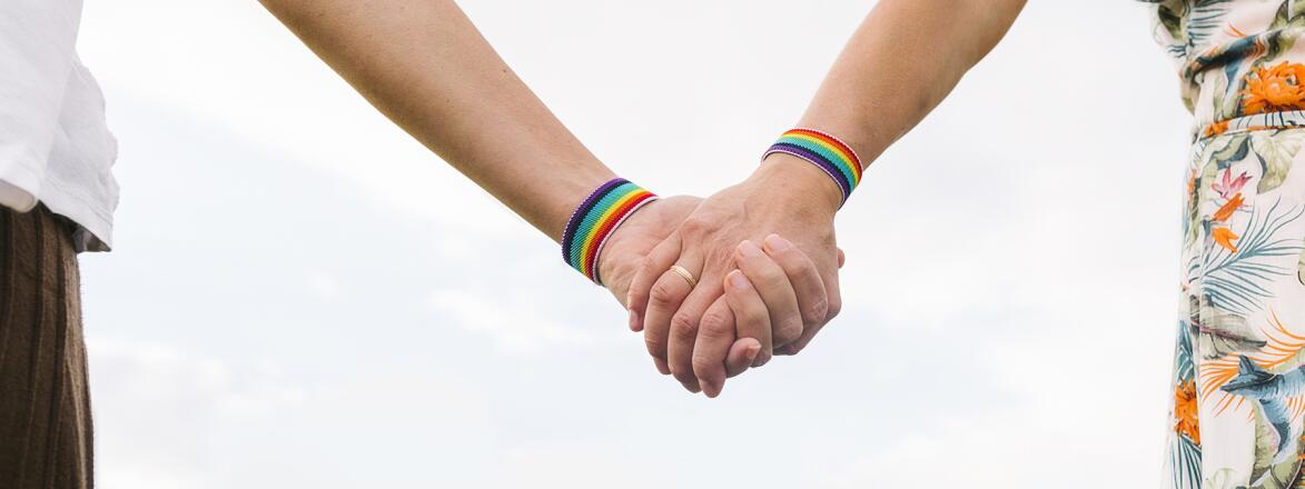 Two women holding hands each wearing a rainbow bracelet and one with a wedding band on