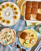 Overhead image of four delicious appetizers including Salmon Dip, Hummus, Deviled Eggs and Ham and Cheese Rolls on a stripe cloth
