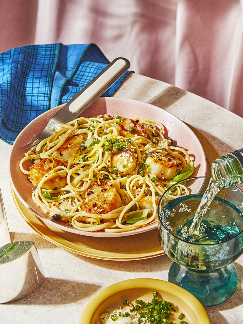 Seared Sea Scallops and pasta with garnish are in a pink bowl with a silver and pink fork. On the white stone surface sits from bottom right to top left: a blue wine glass, a yellow garnish dish, a silver salt shaker and a blue checkered napkin. There is a pale pink curtain coming into frame in the background.