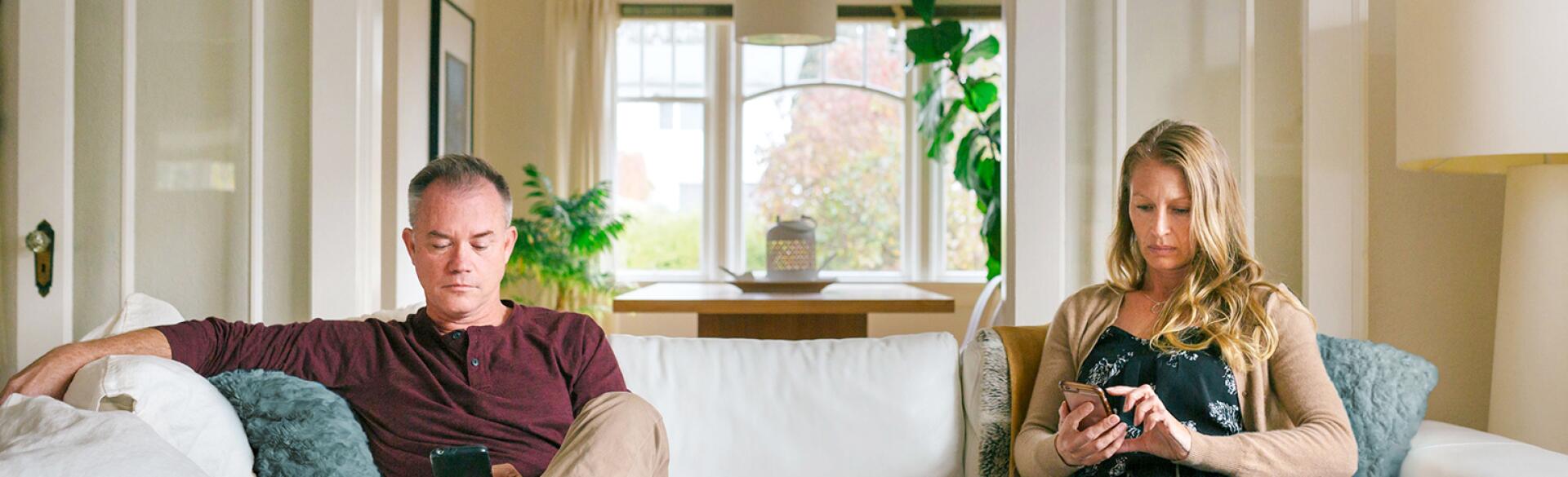 An image of a husband and wife sitting on opposite ends of the couch, distracted from each other by their cellphones.