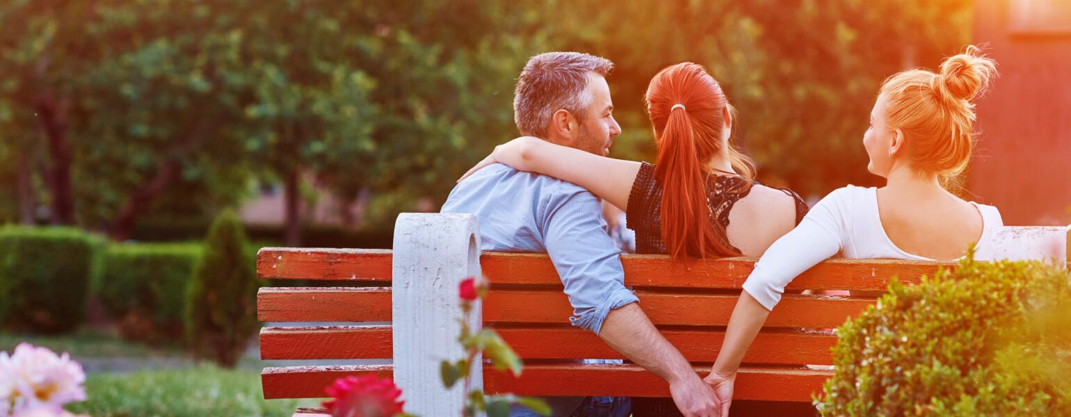 A photo of a husband and wife sitting with a woman on a park bench. While the wife has her arm around her husband's shoulders, he is holding hands with the other woman behind her back.