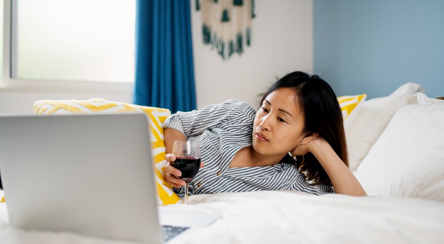 woman watching tv on her computer while laying on her bed with a glass of red wine