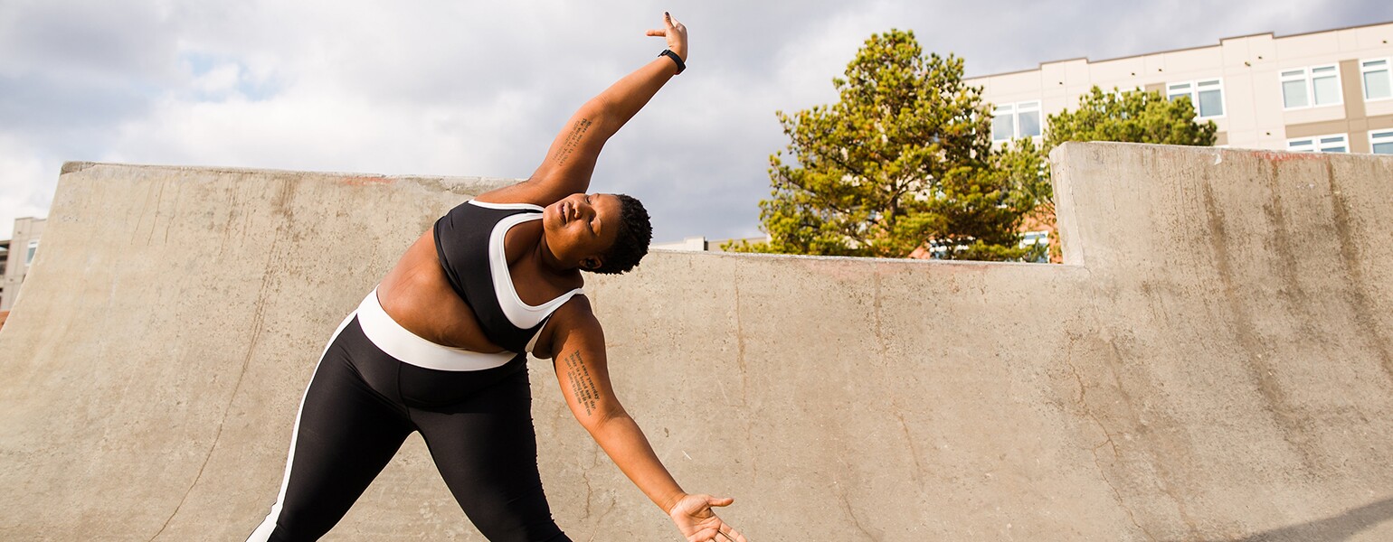 A woman stands on a ramp posing for an Instagram photo, stretching to her left side.