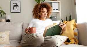 Young multiracial woman reading a book at home, drinking tea sitting on the sofa in cozy living room.