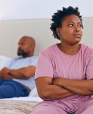 Black couple with arms crossed sitting apart from each other on their bed