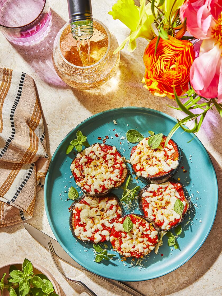 Vegetarian Eggplant Pizzas sit on a teal plate with garnishes on the side. The table is a light stone. The remainder of the table scape has silverware, a patterned brown, white and black napkin, as well as a vase with flowers that are orange, pink and yellow. A pink water tumbler and a glass of wine with at the top left corner. White wine is being poured into the wine glass.