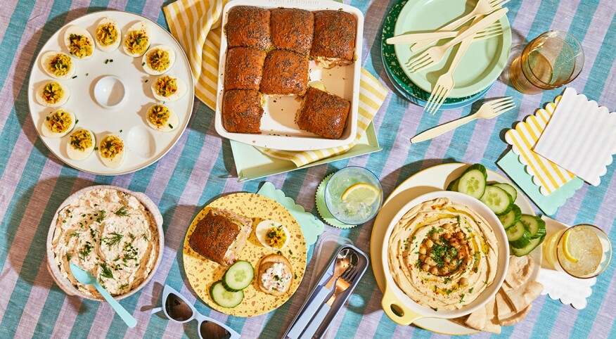 Overhead image of four delicious appetizers including Salmon Dip, Hummus, Deviled Eggs and Ham and Cheese Rolls on a stripe cloth