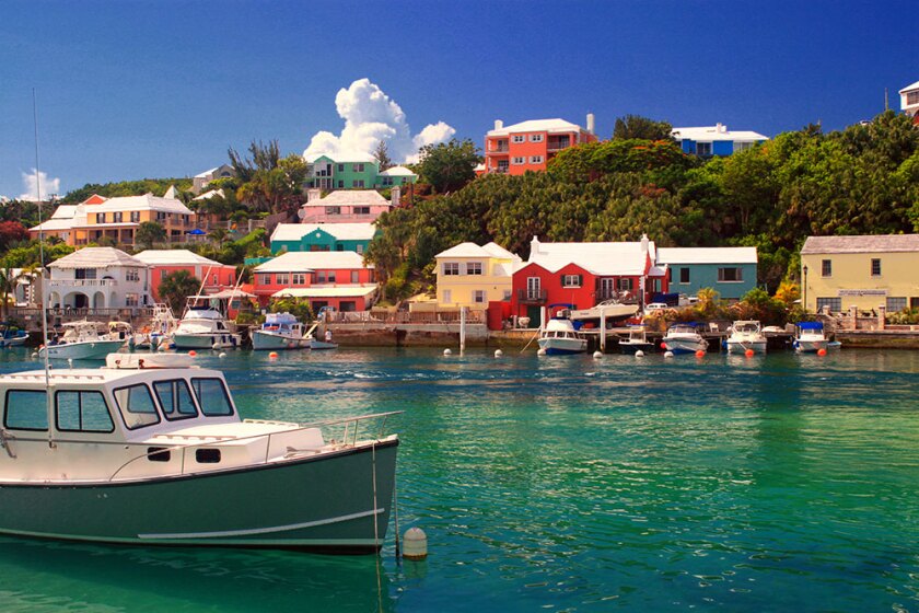 Green and white boat in Bermuda harbor with colorful houses on hill in background.