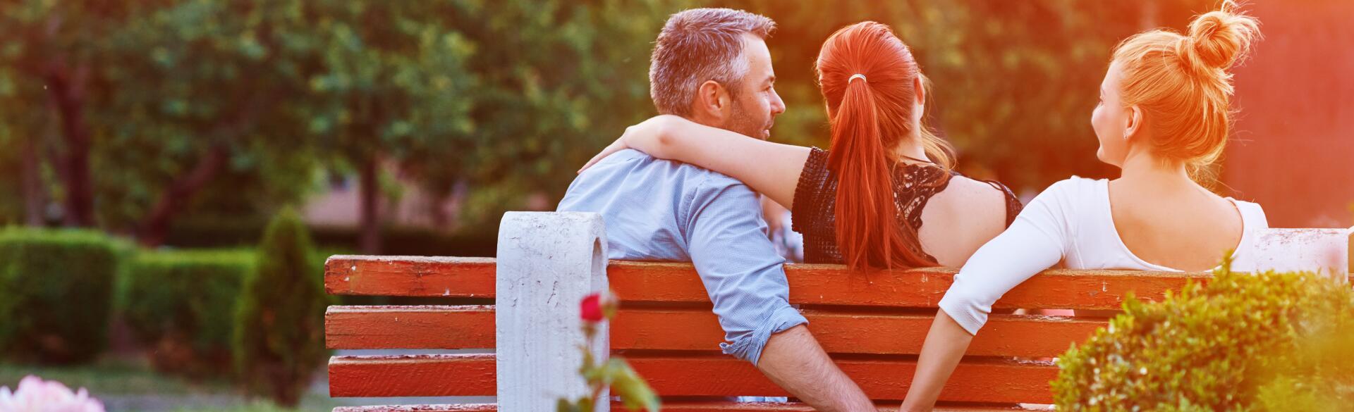 A photo of a husband and wife sitting with a woman on a park bench. While the wife has her arm around her husband's shoulders, he is holding hands with the other woman behind her back.