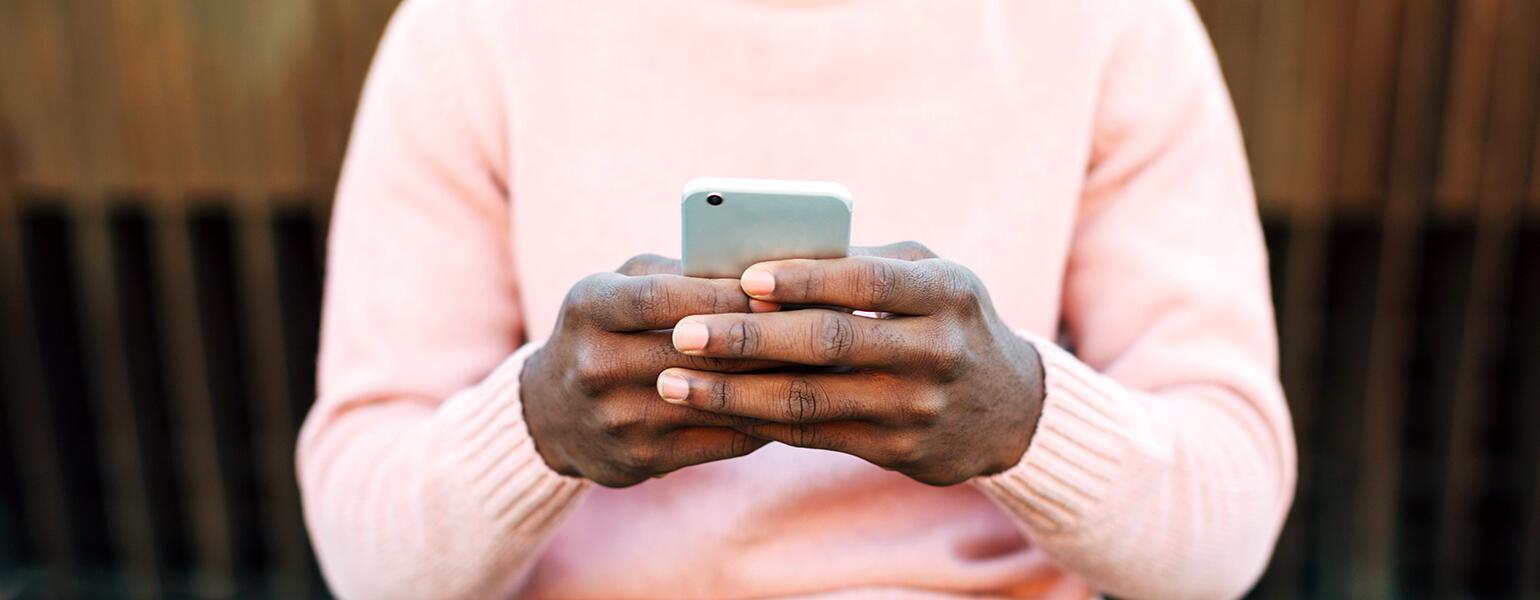 A man holds a cellphone, typing to an unknown person while sitting outside.