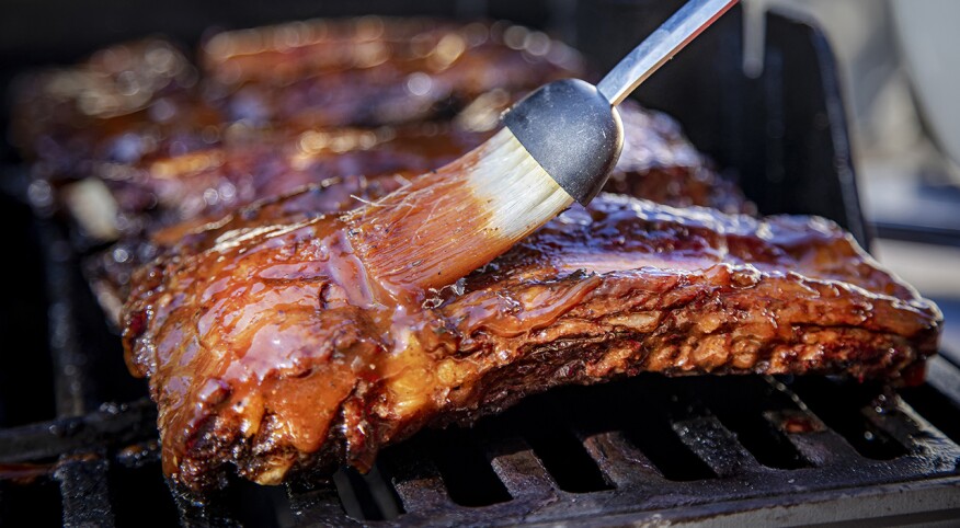 Beef ribs being basted with bbq sauce on a grill with a basting brush 