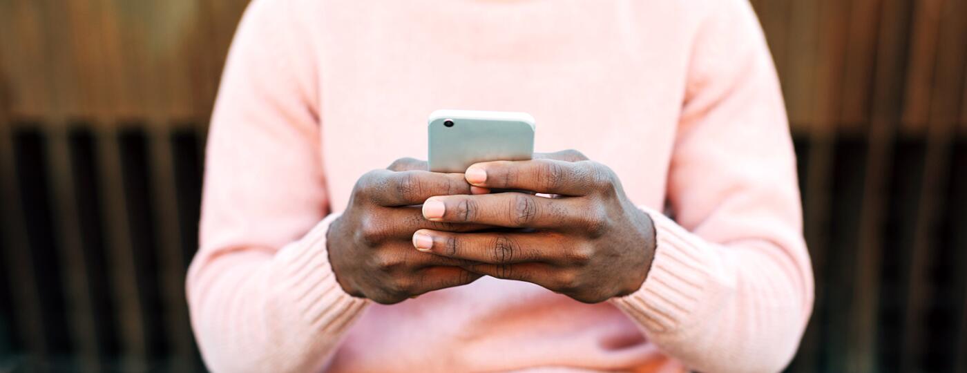 A man holds a cellphone, typing to an unknown person while sitting outside.