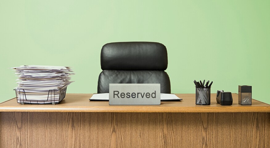 Office desk with overflowing inbox, empty desk chair with a reserved sign in front, pale green wall 