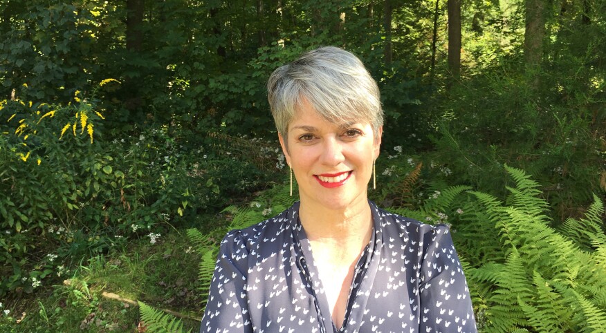 woman with a pixie haircut, grey hair smiles with her arms crossed wearing a blue long sleeve shirt in front of trees