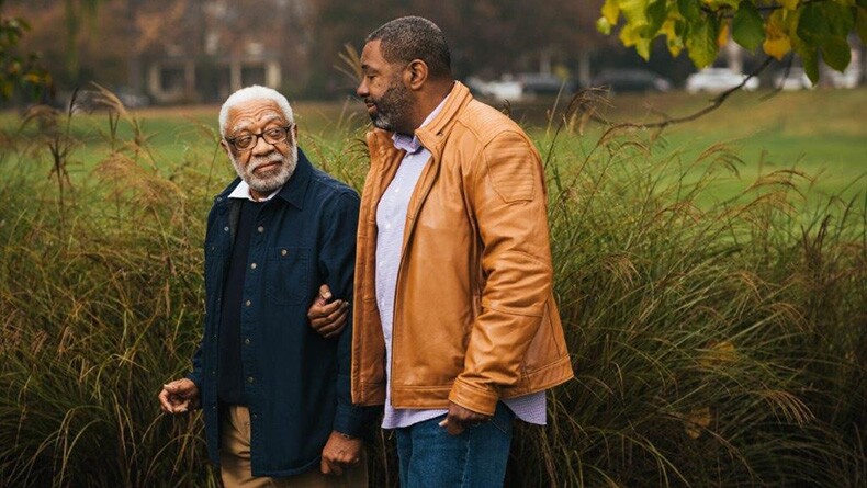 A senior man walks on a trail with his adult son, who is his caregiver.