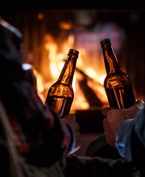 Two men rest with bottles of beer in hand by the fireplace.