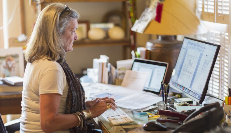 older Caucasian woman sits at a computer with paperwork around her
