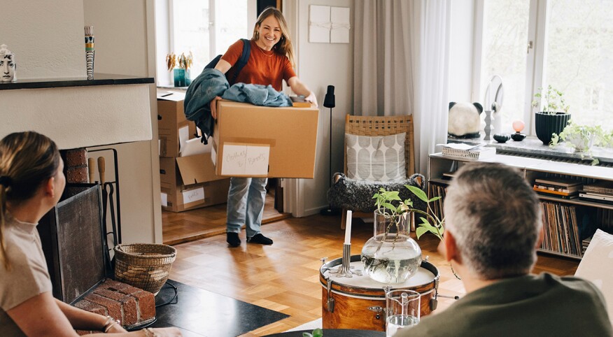 College student returns home with boxes, parents sitting on sofa as she walks in the door