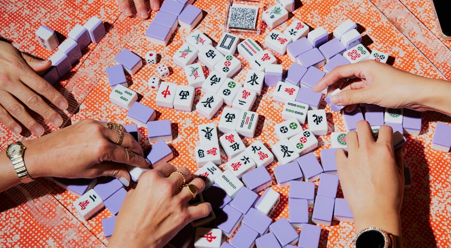 Four women playing Mahjong with violet tiles on a bright orange colored surface