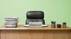 Office desk with overflowing inbox, empty desk chair with a reserved sign in front, pale green wall