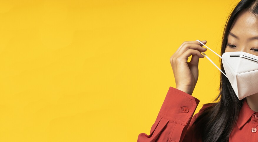 Asian Woman putting on a face mask standing in front of a bright yellow background