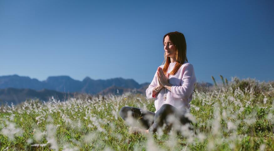 Beautiful Young Woman Meditates In Grassland With Mountains In The Background