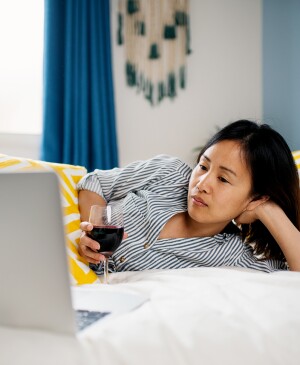 woman watching tv on her computer while laying on her bed with a glass of red wine