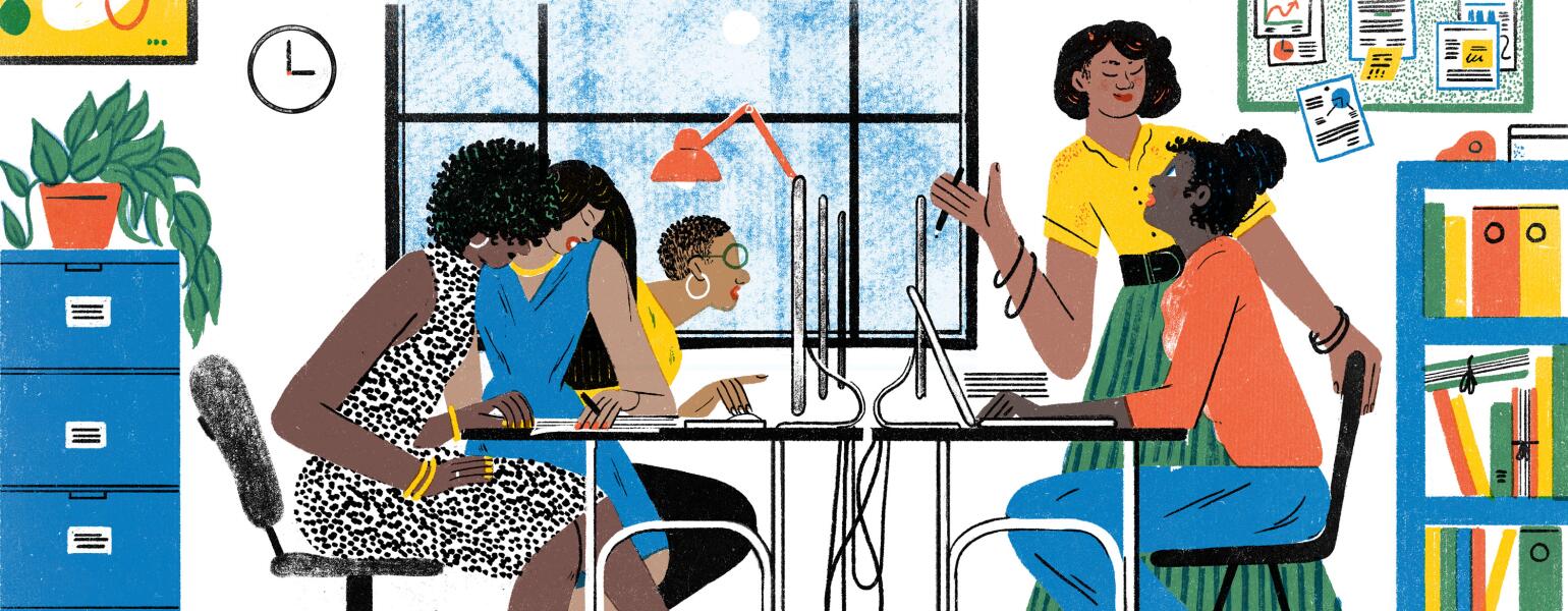 A group of women in an office space sit around a communal desk, talking and working on computers.
