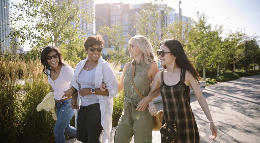 4 women walking down a sunny city sidewalk with linked arms
