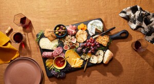 Charcuterie board with a display of cheese and meats on a rust colored table cloth, red wine, plates and napkins
