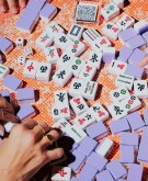 Four women playing Mahjong with violet tiles on a bright orange colored surface