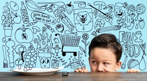 Child hiding his face beneath table peeking over at a plate with drawings of food surrounding it