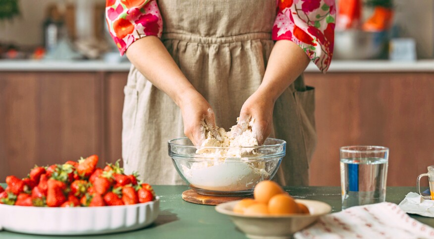 Woman in apron needs dough in a bowl with a large plate of strawberries on the counter close by