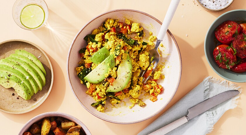 A tofu scramble dish is centered on a peach surface. The tofu scramble is inside of a speckled pink bowl and includes cooked vegetables like spinach and peppers as well as fresh avocado. To the right of the dish sits, from top, a bowl of salt and spices, and below that a blue bowl with seasoned cherry tomatoes and below that a blue napkin folded with a knife sitting on top. To the left of the dish, from the top, sits a clear glass of sparkling water with a lime round in it, then below that a tan plate with slices of avocado, seasoned, and below that a pink bowl with cooked and seasoned potatoes is half in frame.