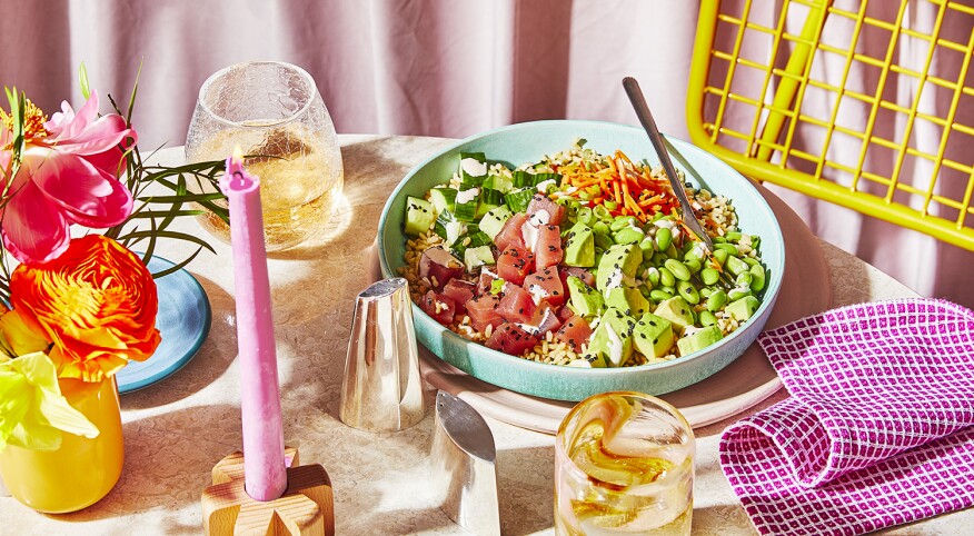 A light stone circular table set for one person. A yellow chair, blue plate, pink checkered napkin and pink candle that's lit sit on the table. As well as a yellow vase filled with yellow, orange and pink flowers. A silver set of salt and pepper shakers, a wine glass and a water tumbler sit off to the side of the plate. Tuna Poke bowl with fresh tuna, avocado, edamame, and carrots sit inside the plate.