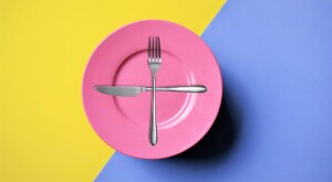 pink plate, silver fork, silver knife, plus sign on plate, yellow blue background, photograph