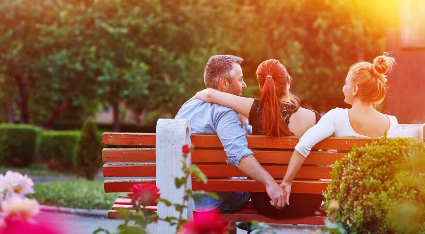 A photo of a husband and wife sitting with a woman on a park bench. While the wife has her arm around her husband's shoulders, he is holding hands with the other woman behind her back.