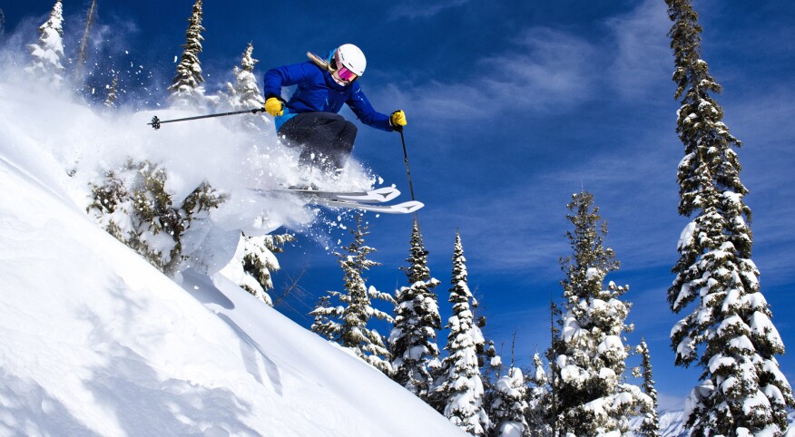 woman skiing at sun valley catching air over a hill with snow covered trees behind her