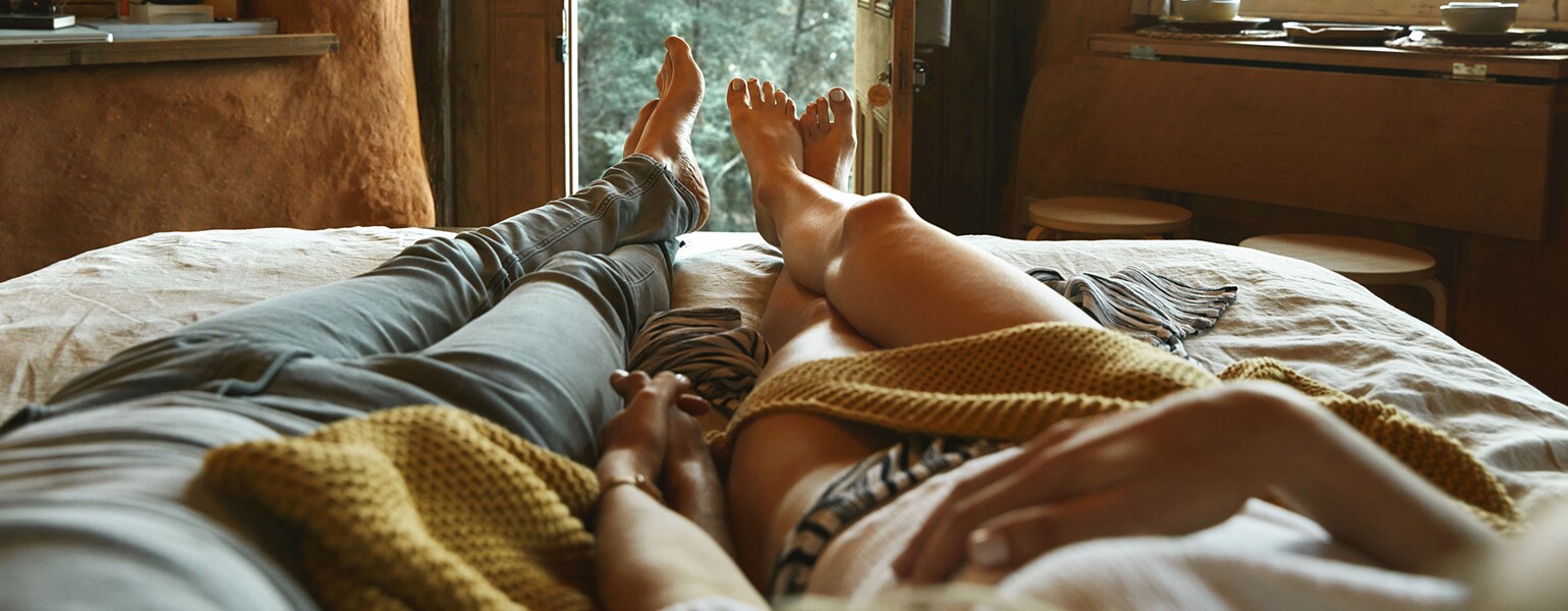 An image of a man and women's legs in bed, where they are lounging together and holding hands.