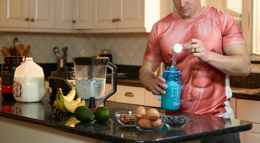 Man in kitchen wearing a muscle t-shirt and pouring protein powder into shaker