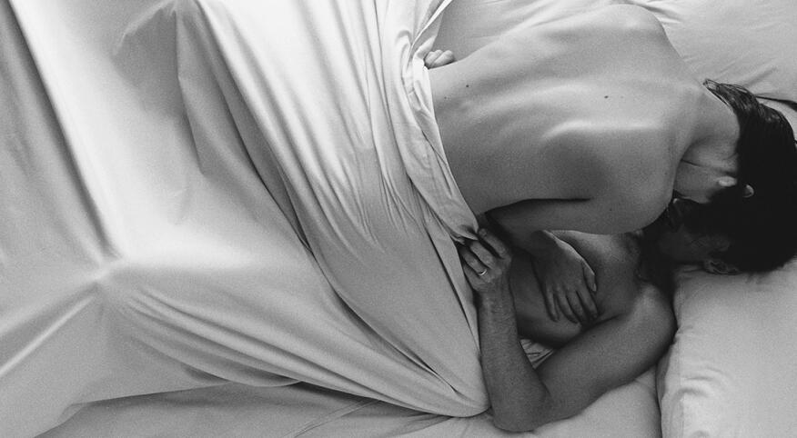 A naked woman lays on top of a man, making love in bed under a white sheet.